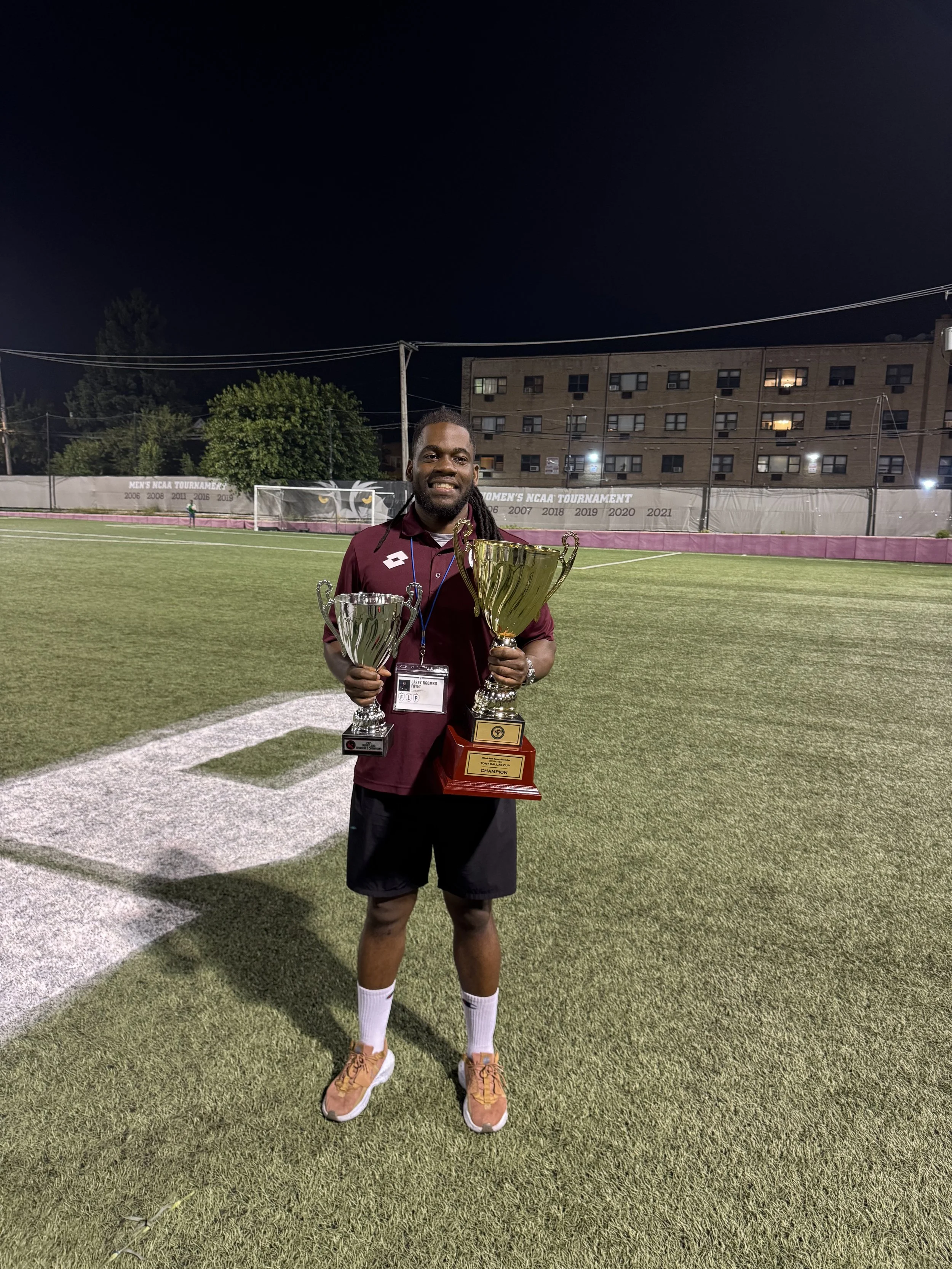 A man standing on a football field at night holding two large trophies, smiling, with a building and trees in the background.