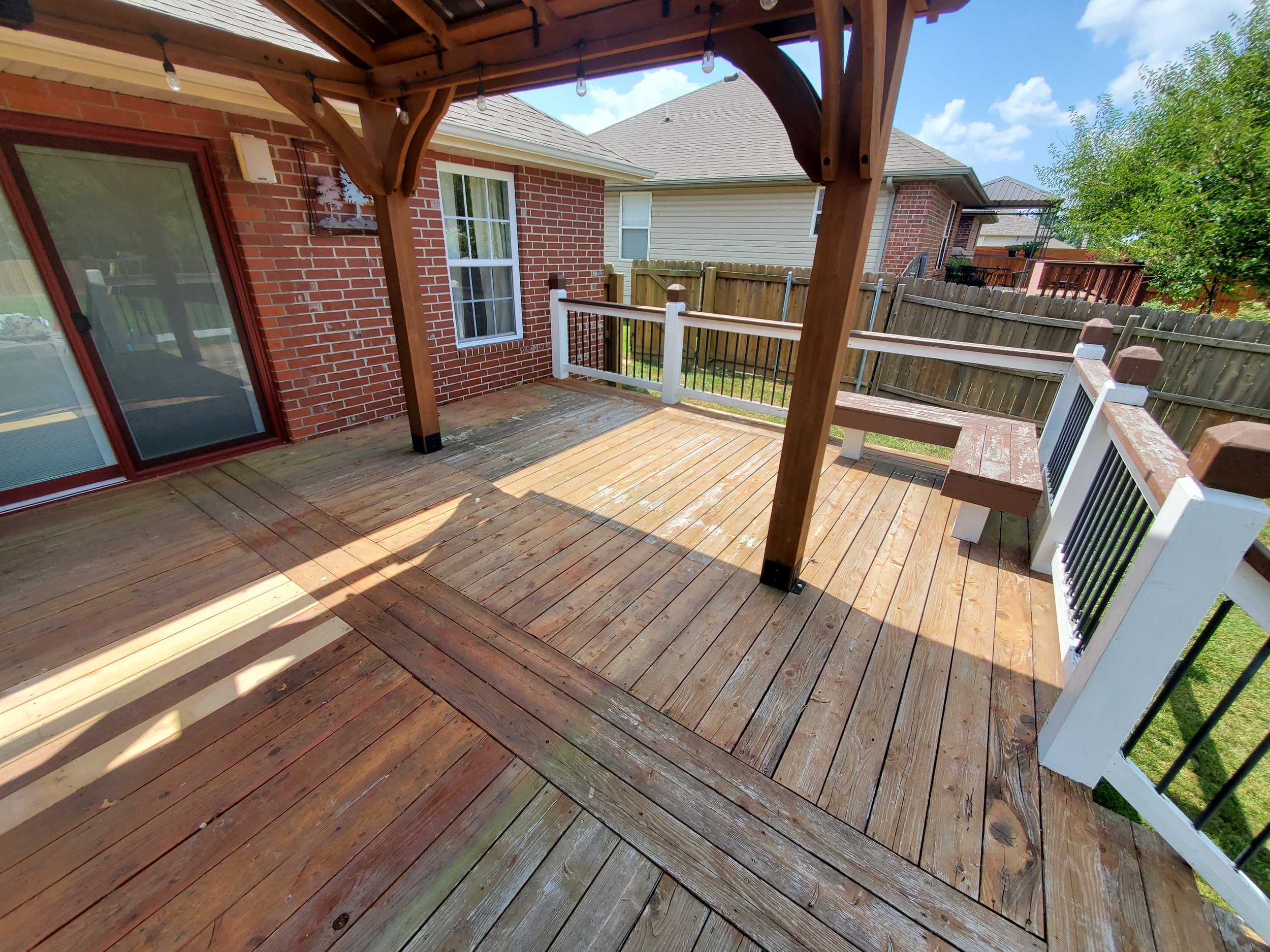 Wooden outdoor deck with a seating bench, surrounded by a white and black railing, attached to a brick house with sliding glass door and window, bathed in sunlight under a partly cloudy sky.