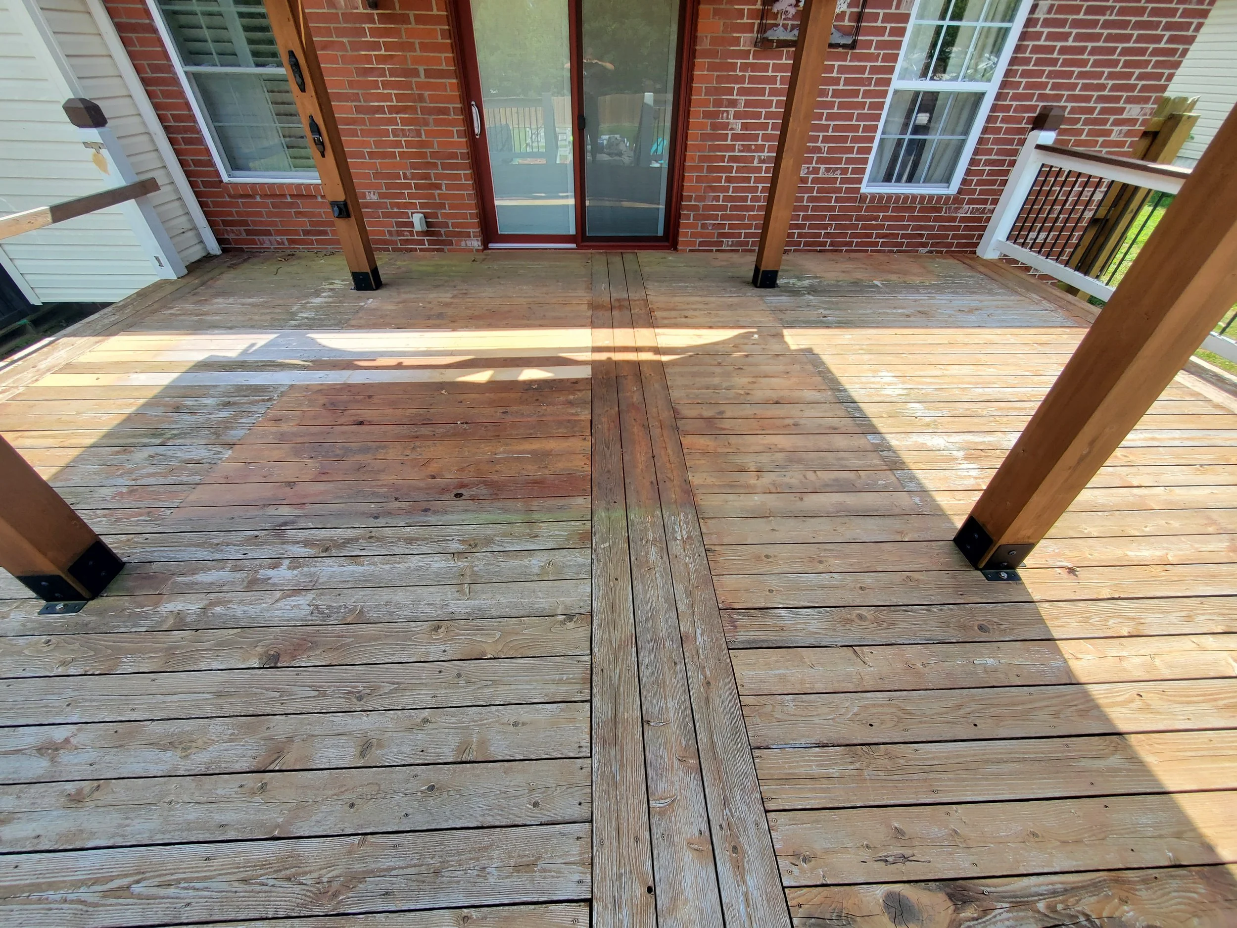 View of a newly built wooden deck attached to a brick house with a sliding glass door, with supporting posts and railing partially visible, and sunlight casting shadows on the deck.