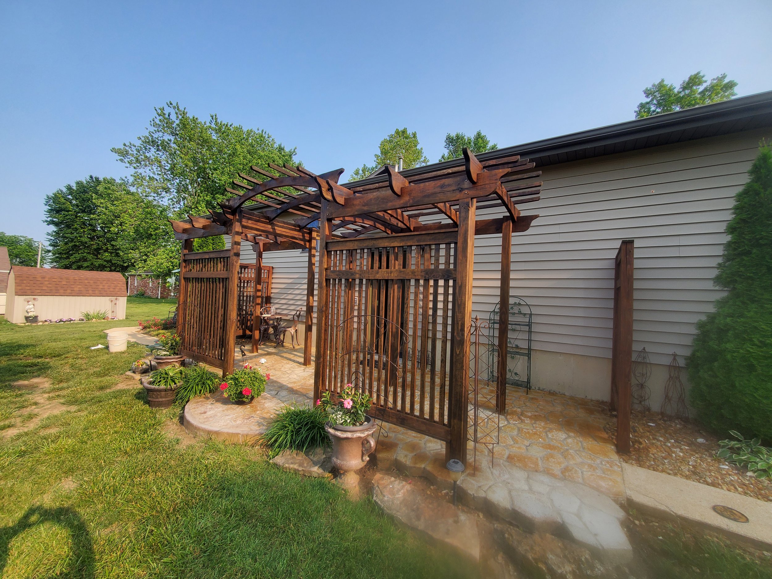 Backyard patio with a wooden pergola, patio table and chairs, potted plants, and a grassy lawn, next to a house with white siding.