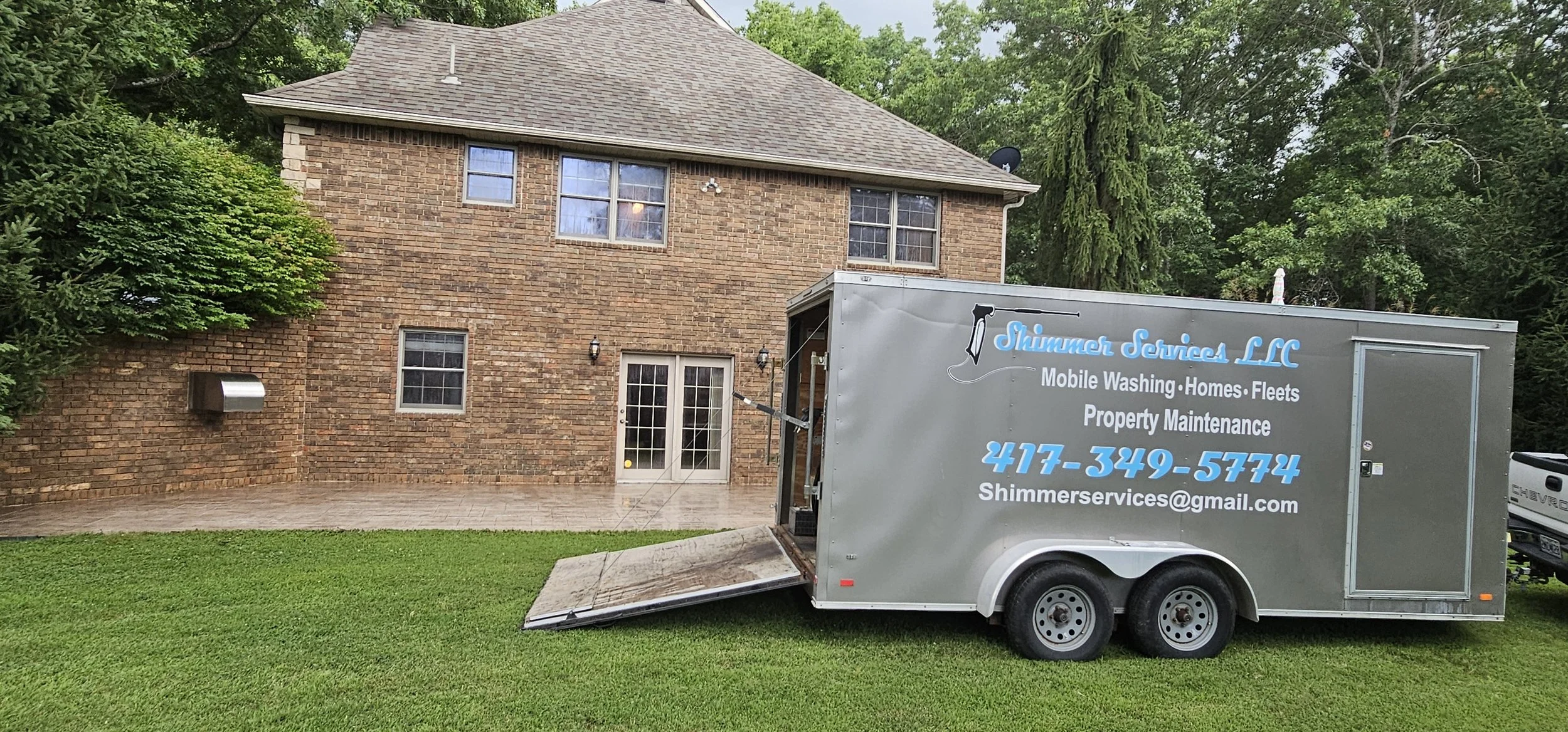 A mobile washing and property maintenance trailer parked on a well-maintained lawn in front of a brick house with multiple windows, surrounded by green trees.
