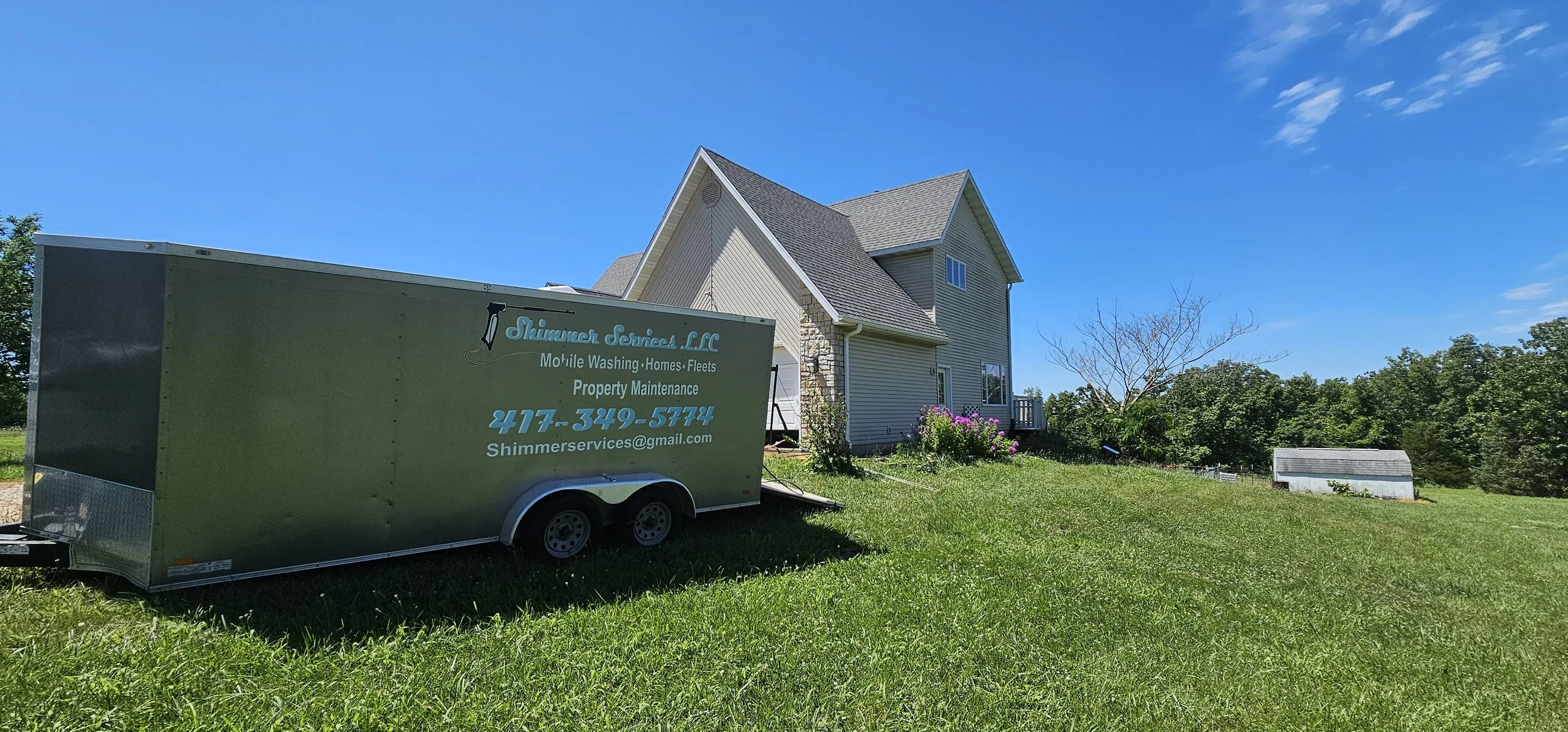 Lawn with mobile washing trailer parked near suburban house with flower garden, leafless tree, and small white shed under a blue sky.
