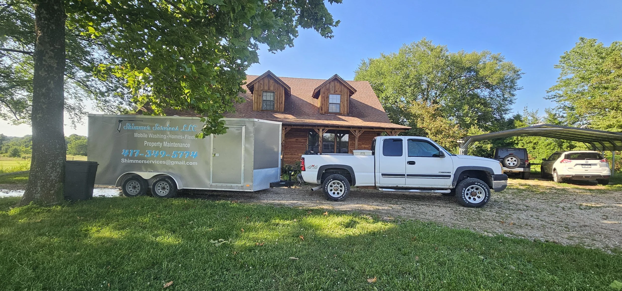 A white pickup truck with a trailer attached parked in front of a two-story wooden house with a gabled roof. The trailer has the logo and contact information for Shimmer Services, LLC, a cleaning and property maintenance company. There are two cars p