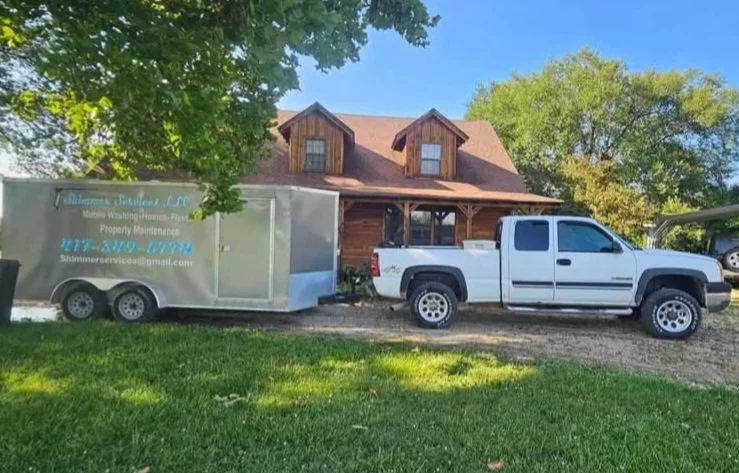 White pickup truck with a trailer attached parked on a grassy area in front of a wooden house, with trees and a blue sky in the background.