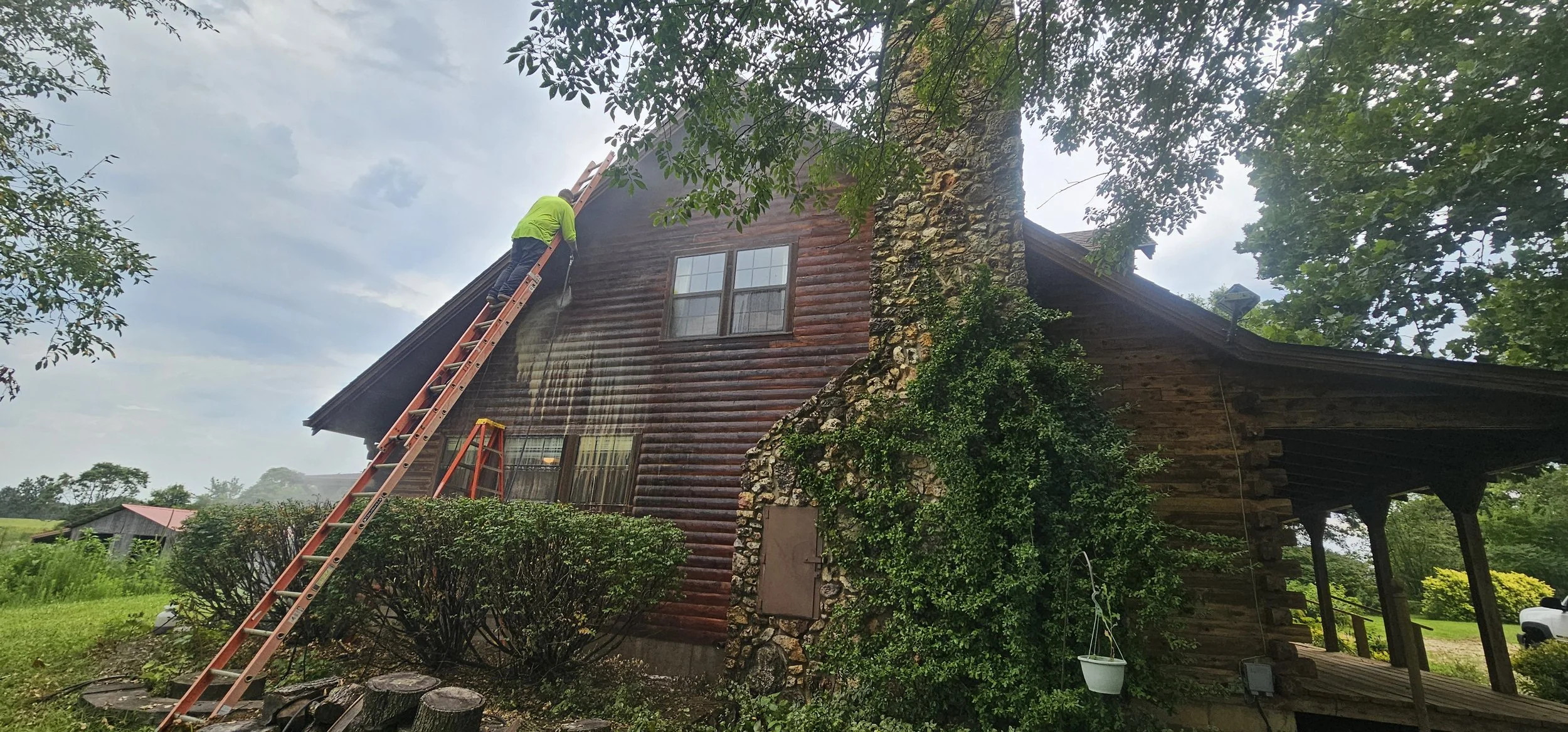 A person in a green shirt and blue jeans on a ladder cleaning the upper windows of a rustic log cabin, surrounded by lush green trees and bushes.