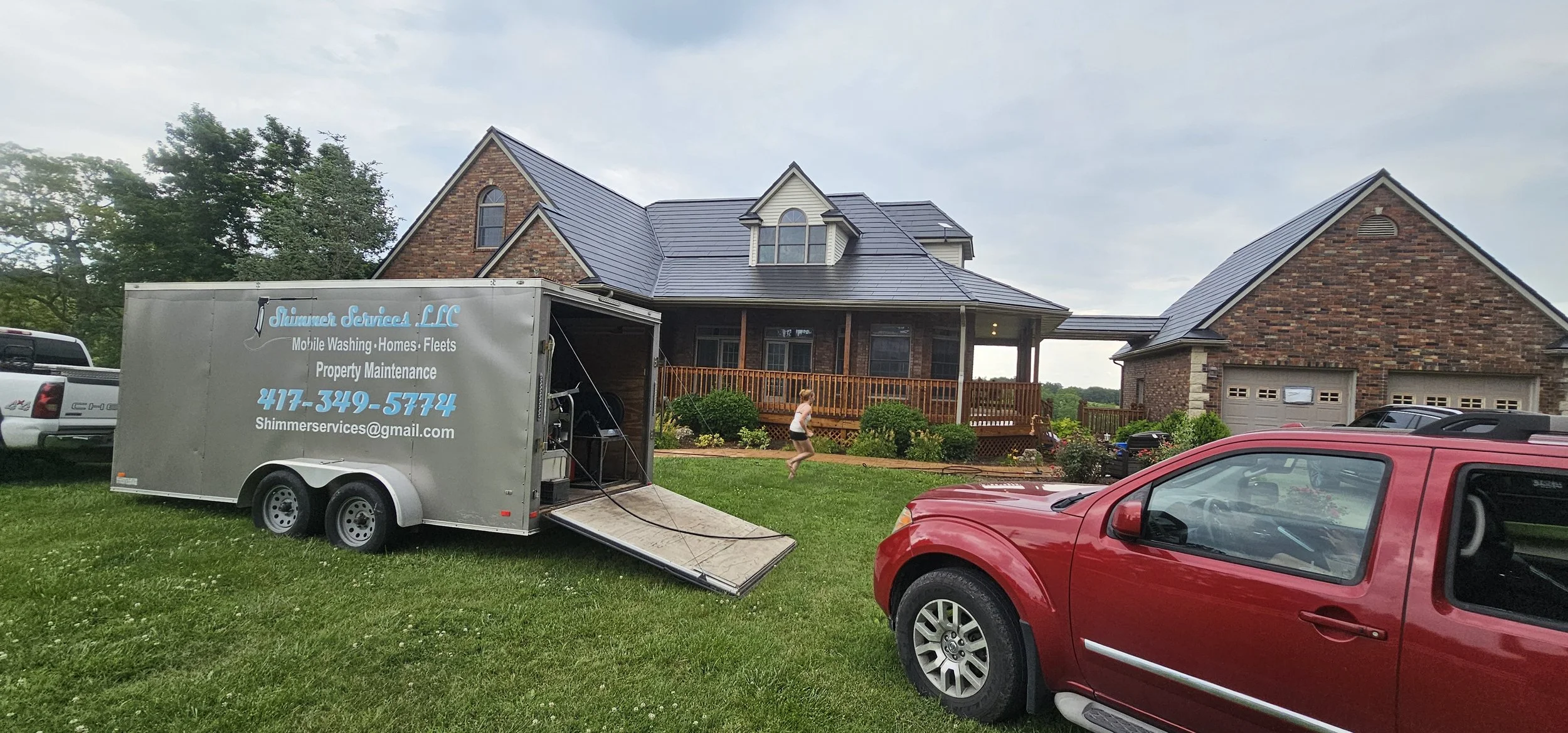 A mobile cleaning service trailer parked on a lawn in front of a brick house with a wooden porch. A person is running across the yard. A red vehicle is parked nearby.