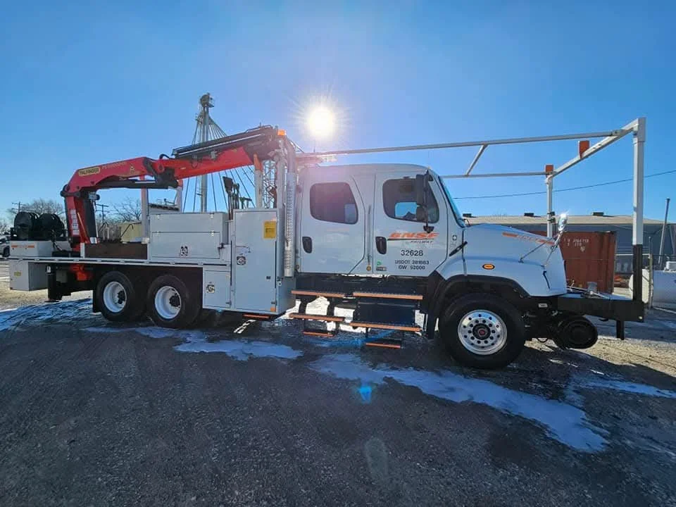 White utility truck with a red crane and metal framework, parked on a gravel lot with snow patches, under a bright sun in a clear blue sky.