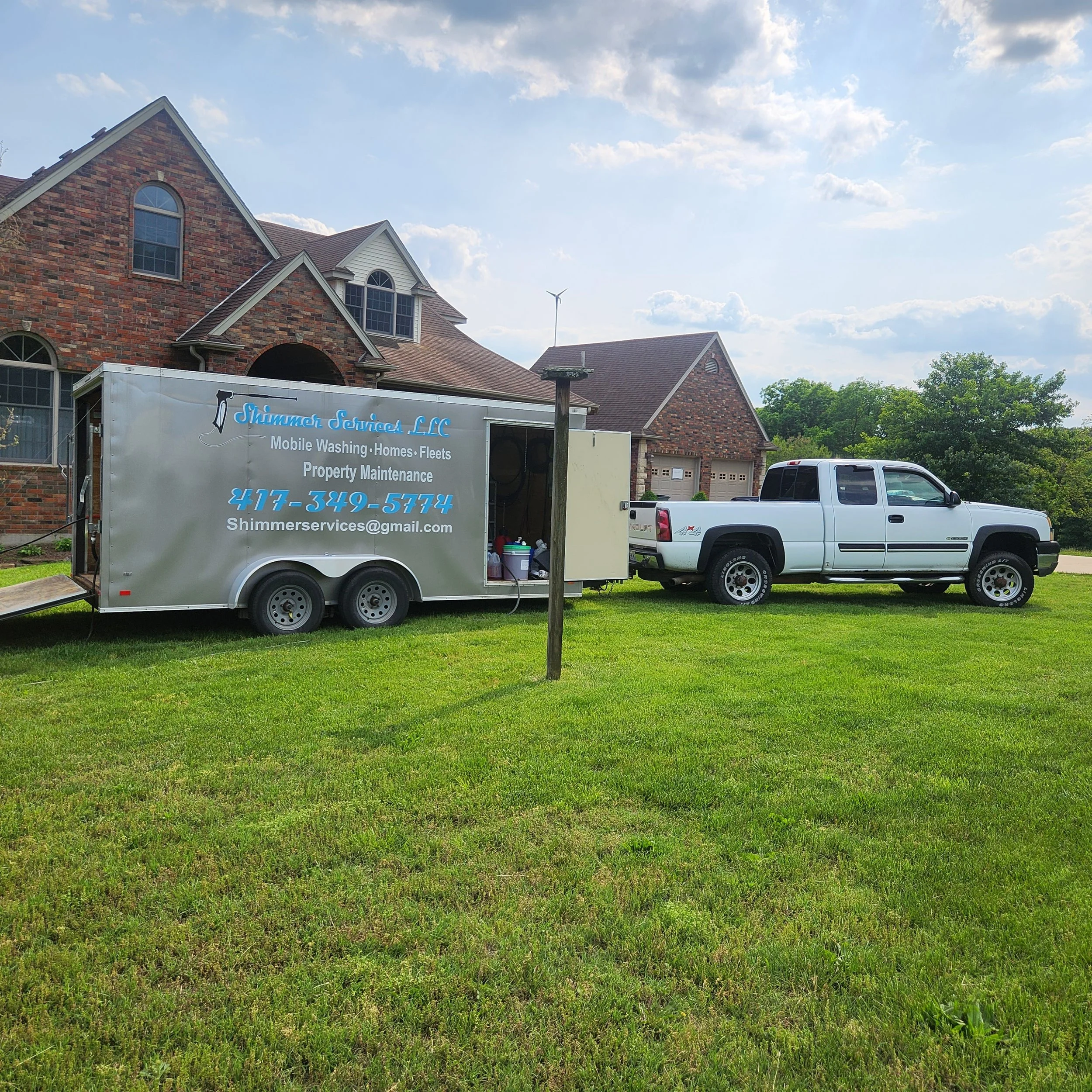 A white pickup truck parked on a lawn with a mobile cleaning trailer attached, in front of a brick house with a garage. The trailer displays the business nameShimmer Services LLC, and contact information.