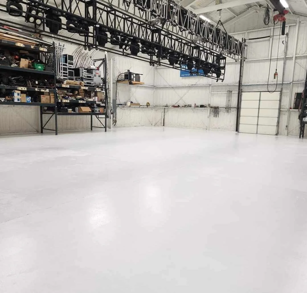 Empty white studio or warehouse with shelves, tools, and equipment along the walls, and large metal light rig hanging from the ceiling.