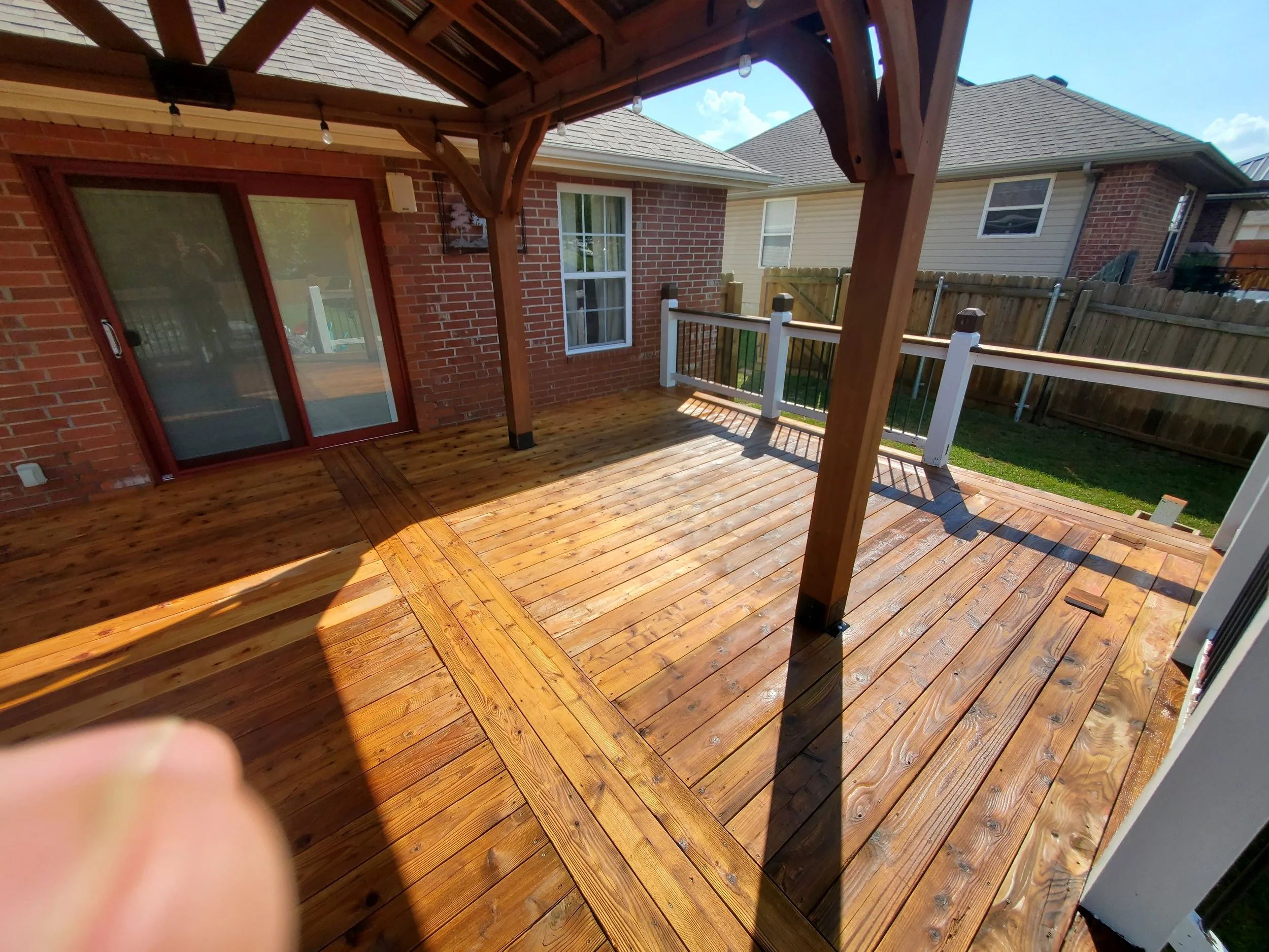 Freshly stained wooden backyard deck with a pergola, outside of a brick house with sliding glass door and windows, surrounded by a wooden fence.