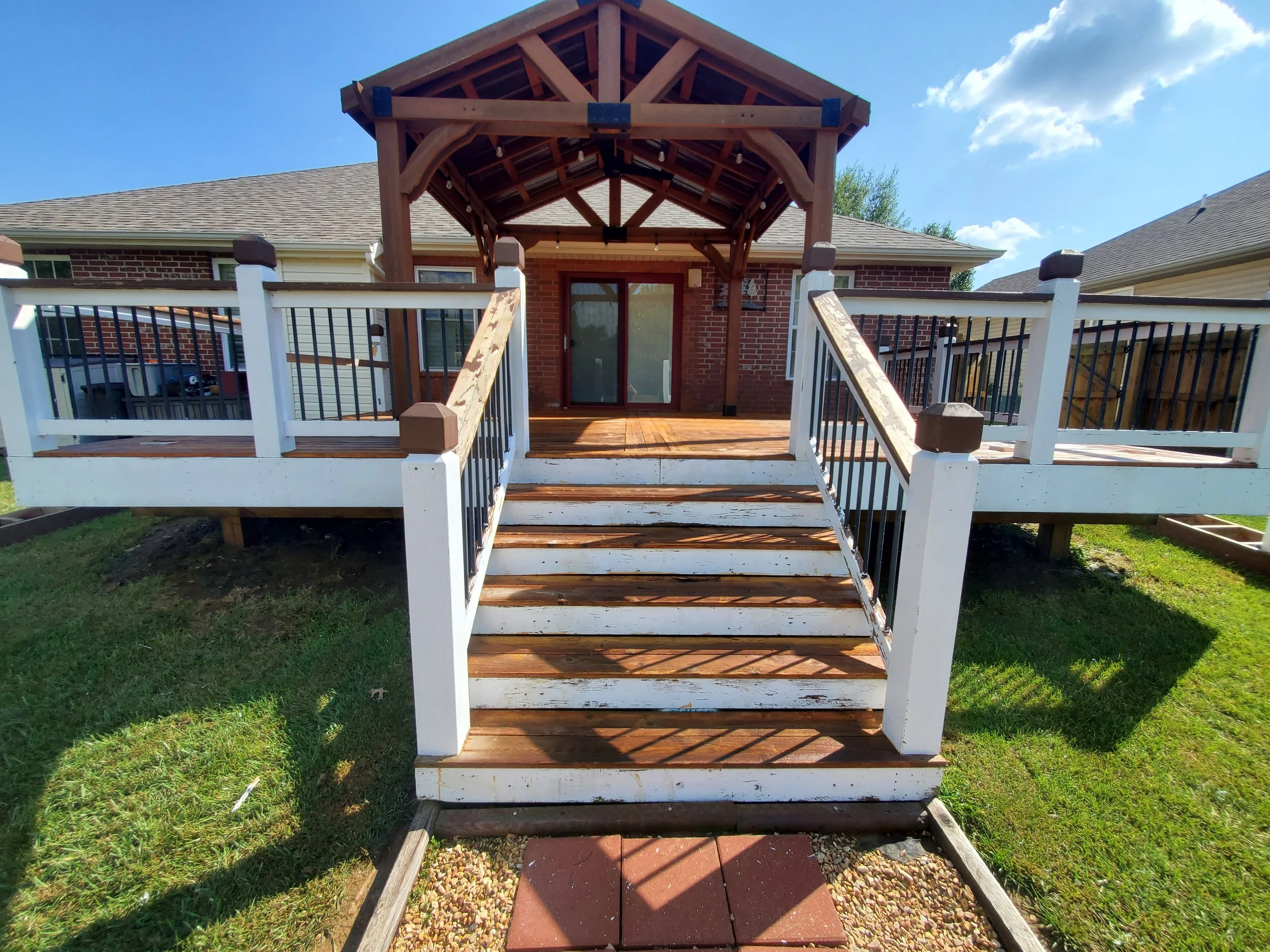 Backyard view of a brick house with a raised wooden deck, featuring white and black railing, a wooden pergola over the deck, and a sliding glass door leading inside.