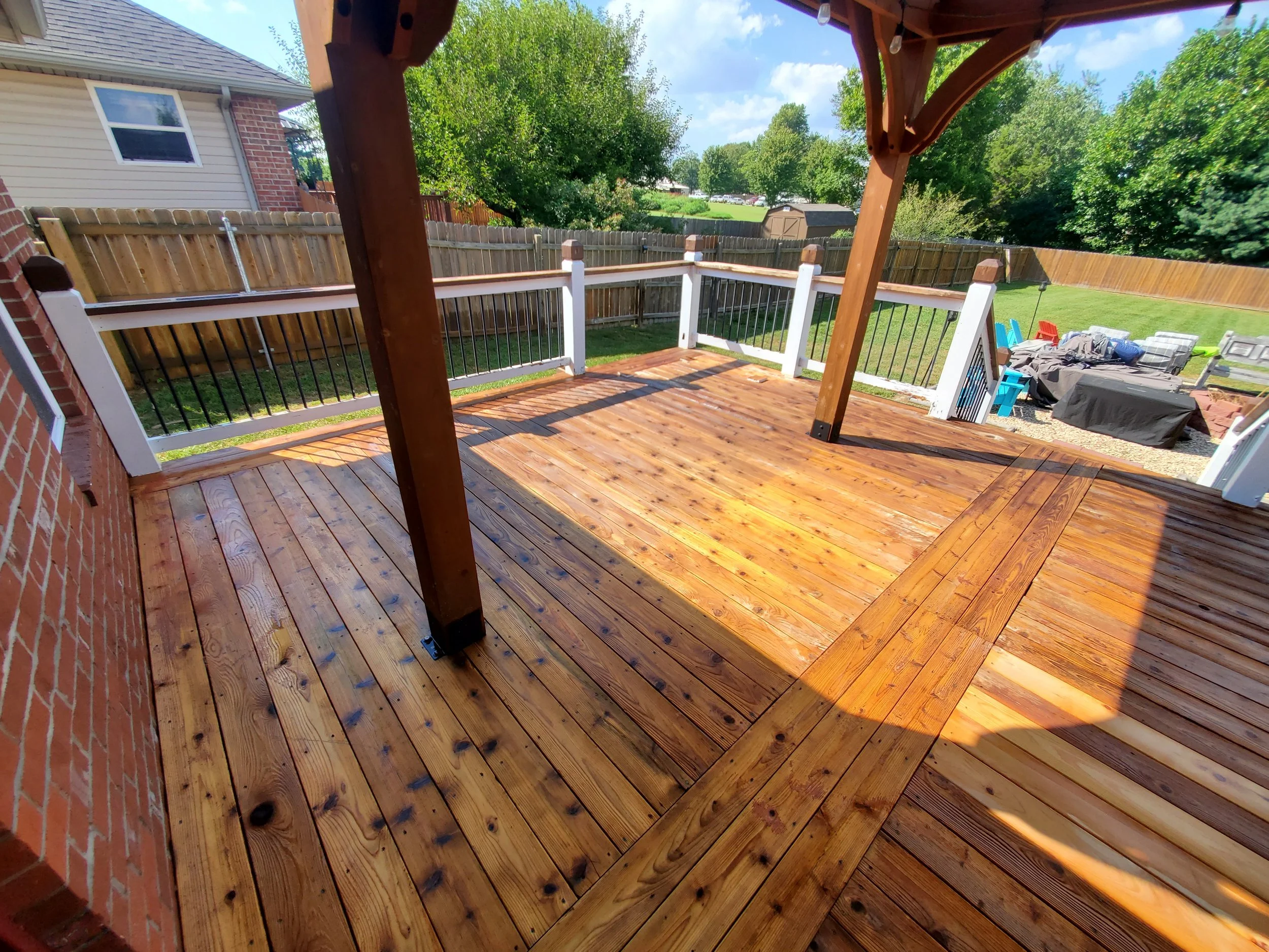 Newly built wooden deck with railing in backyard, partially shaded, with outdoor furniture and a grassy yard enclosed by a fence.