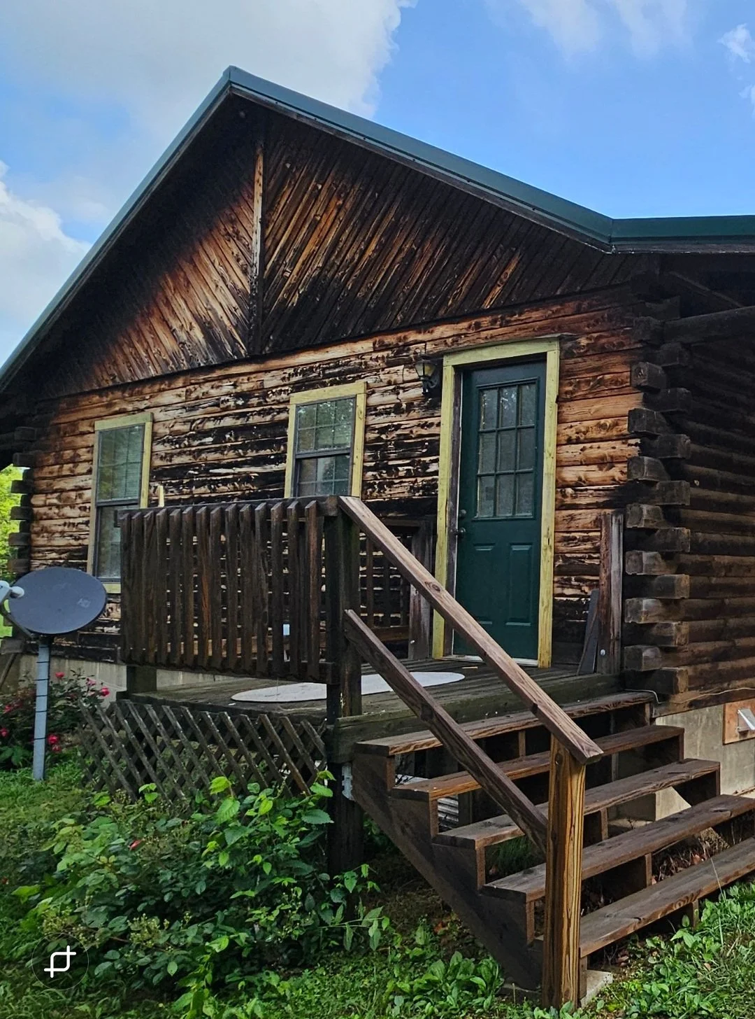 A small wooden house with a green door and two windows, having an elevated porch and stairs, surrounded by green plants and bushes, with a bright blue sky and some clouds in the background.