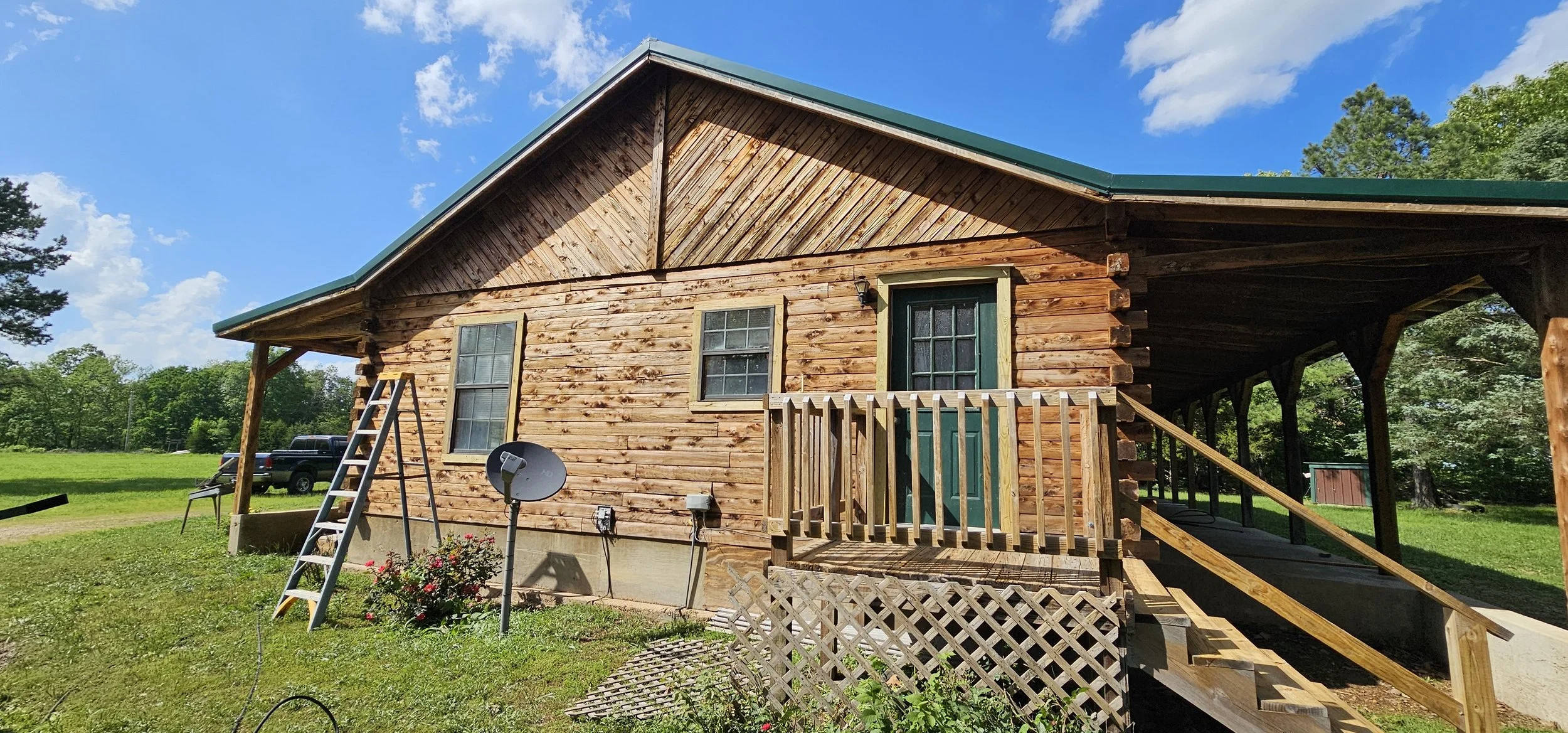 A rustic wooden house with a metal roof, surrounded by a lush green lawn and trees under a blue sky with a few clouds.