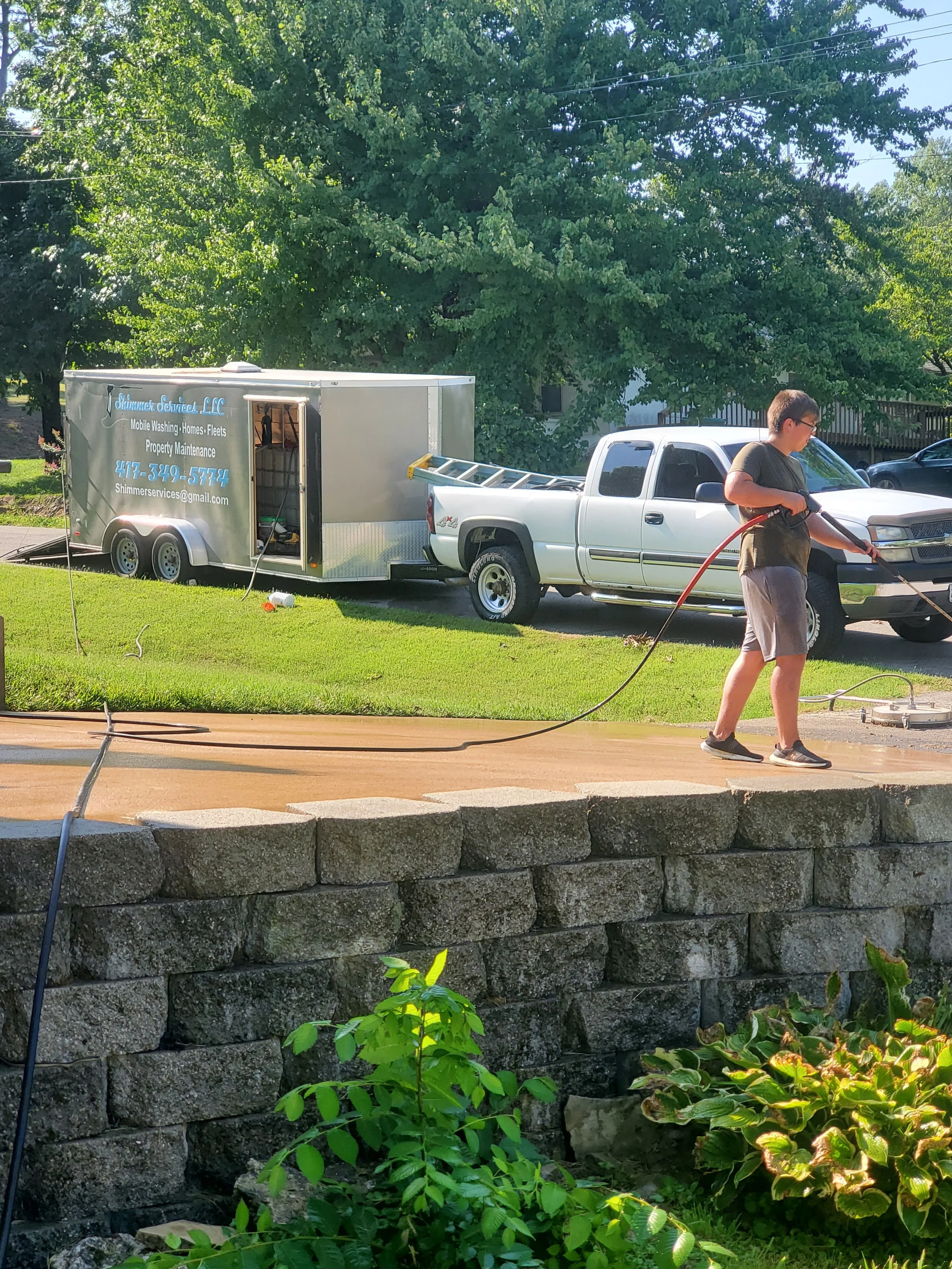 A person pressure-washing a sidewalk in a residential yard with a trailer in the background, trees, green grass, and stone landscaping.