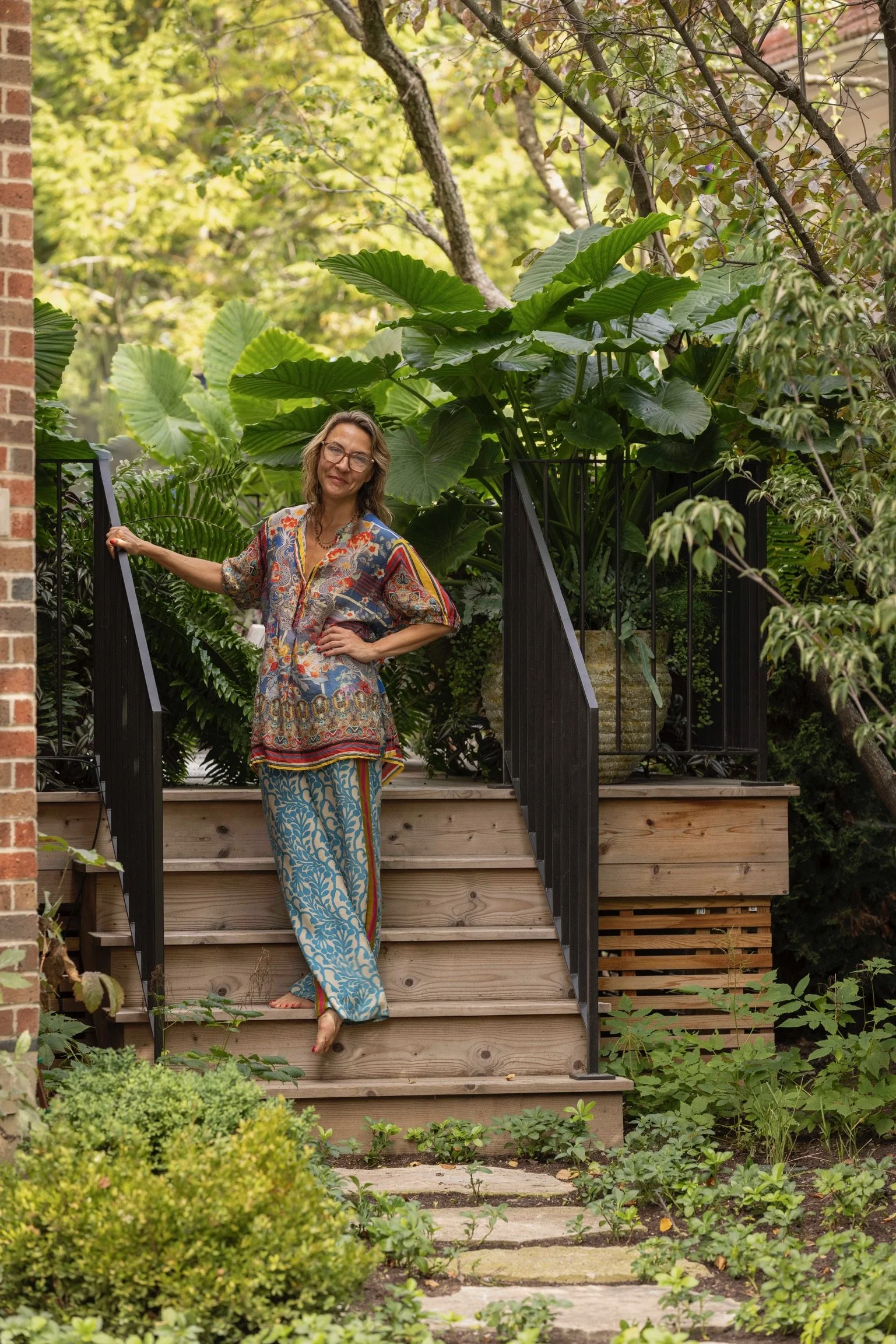 Paige wearing glasses and a long, colorful, patterned dress standing on a wooden deck with large green leaves behind her, surrounded by lush garden plants.