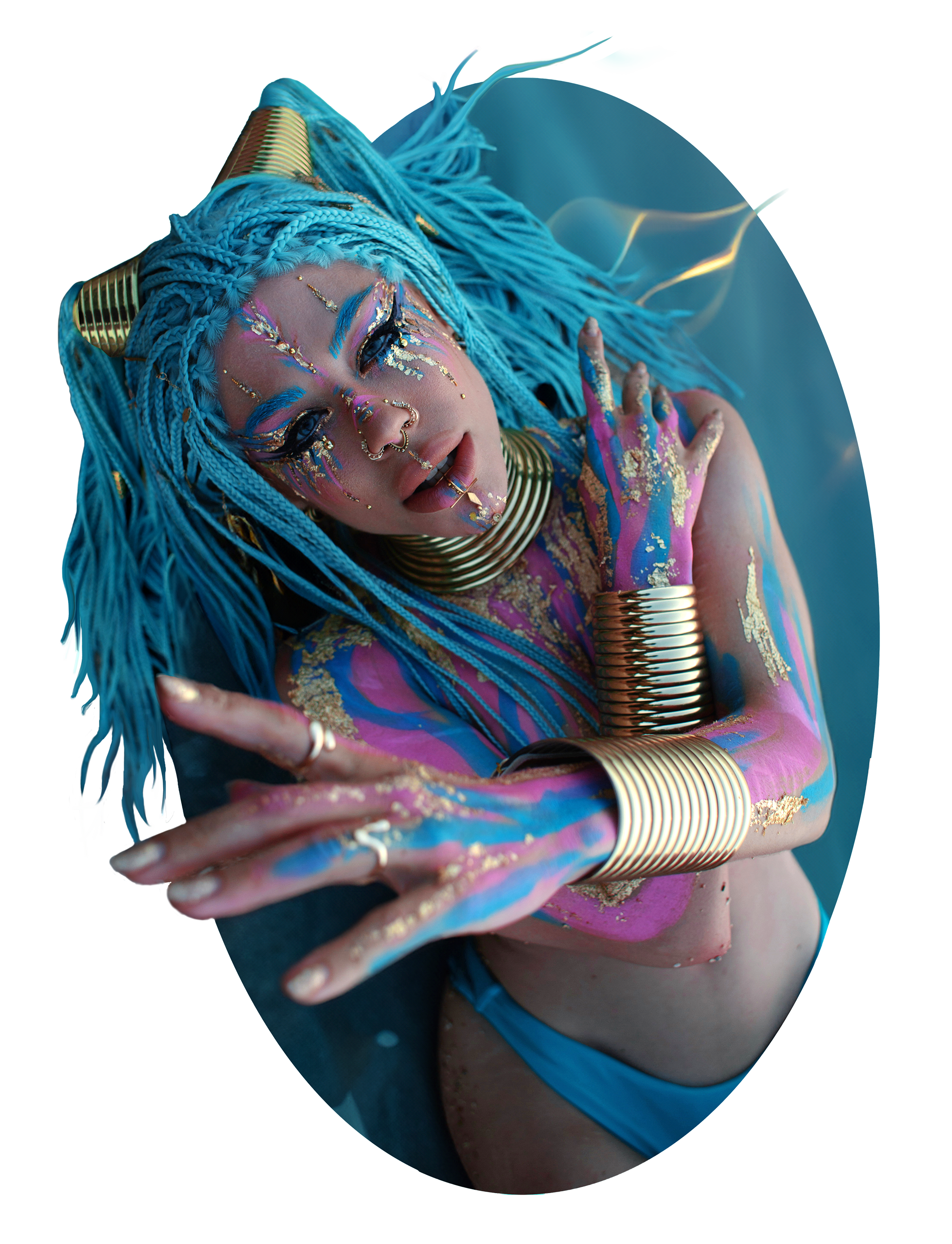Woman with blue dreadlocks, adorned with gold jewelry, colorful makeup, and gold paint on her body, posed with hands near her face.