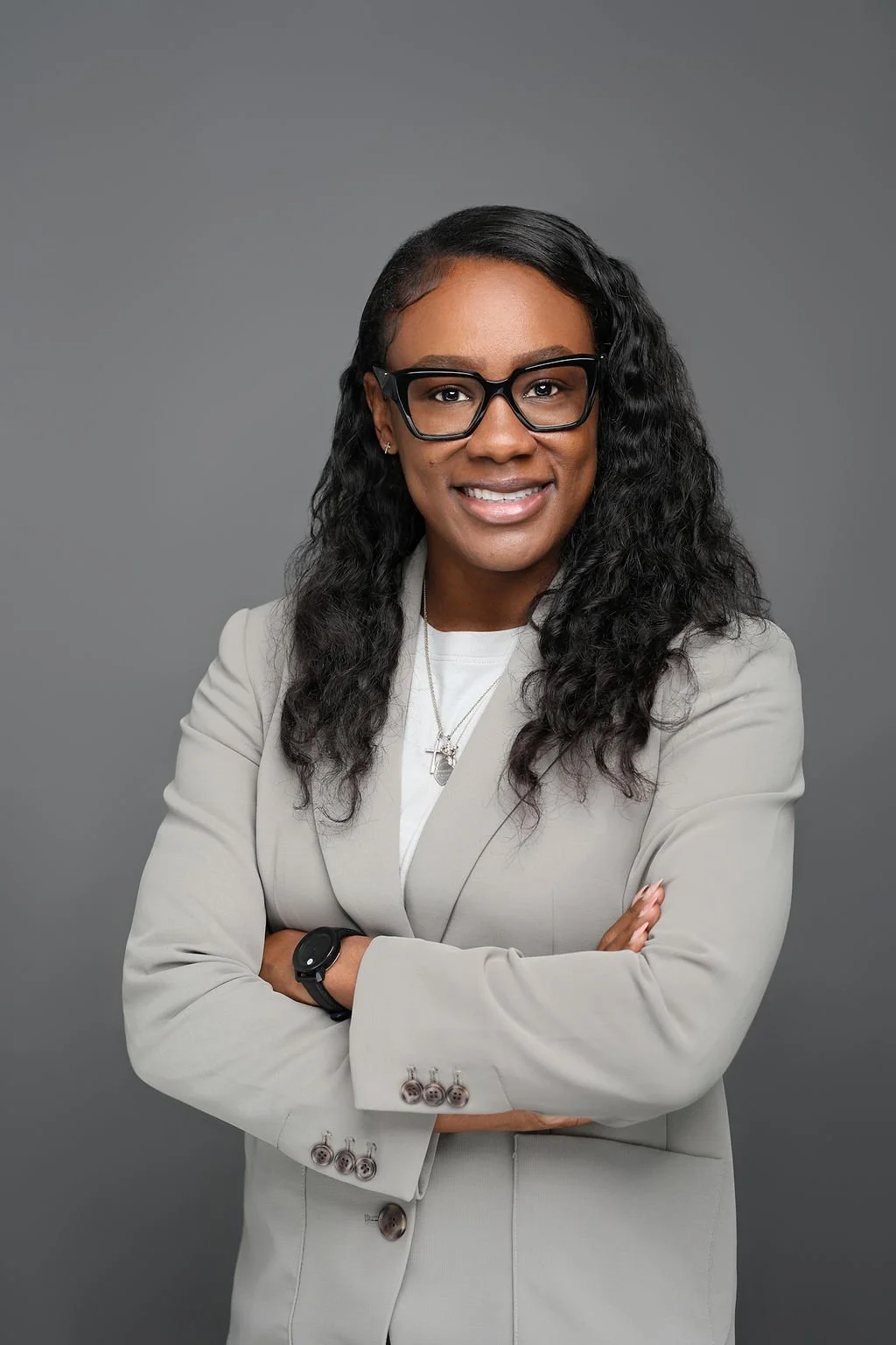 Portrait of Shamiea Green wearing glasses, a light-colored blazer, and jewelry, smiling with arms crossed against a gray background.