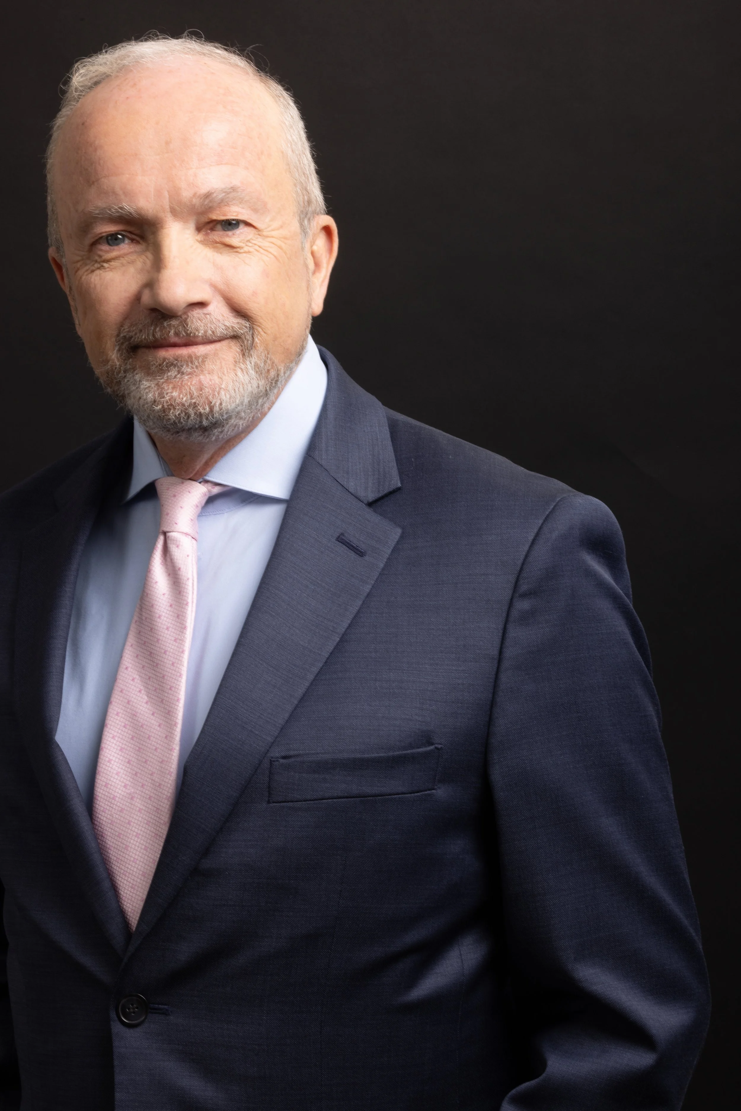 A middle-aged man with gray hair and a beard, wearing a dark suit, light pink tie, and a light blue shirt, smiling softly against a dark background.