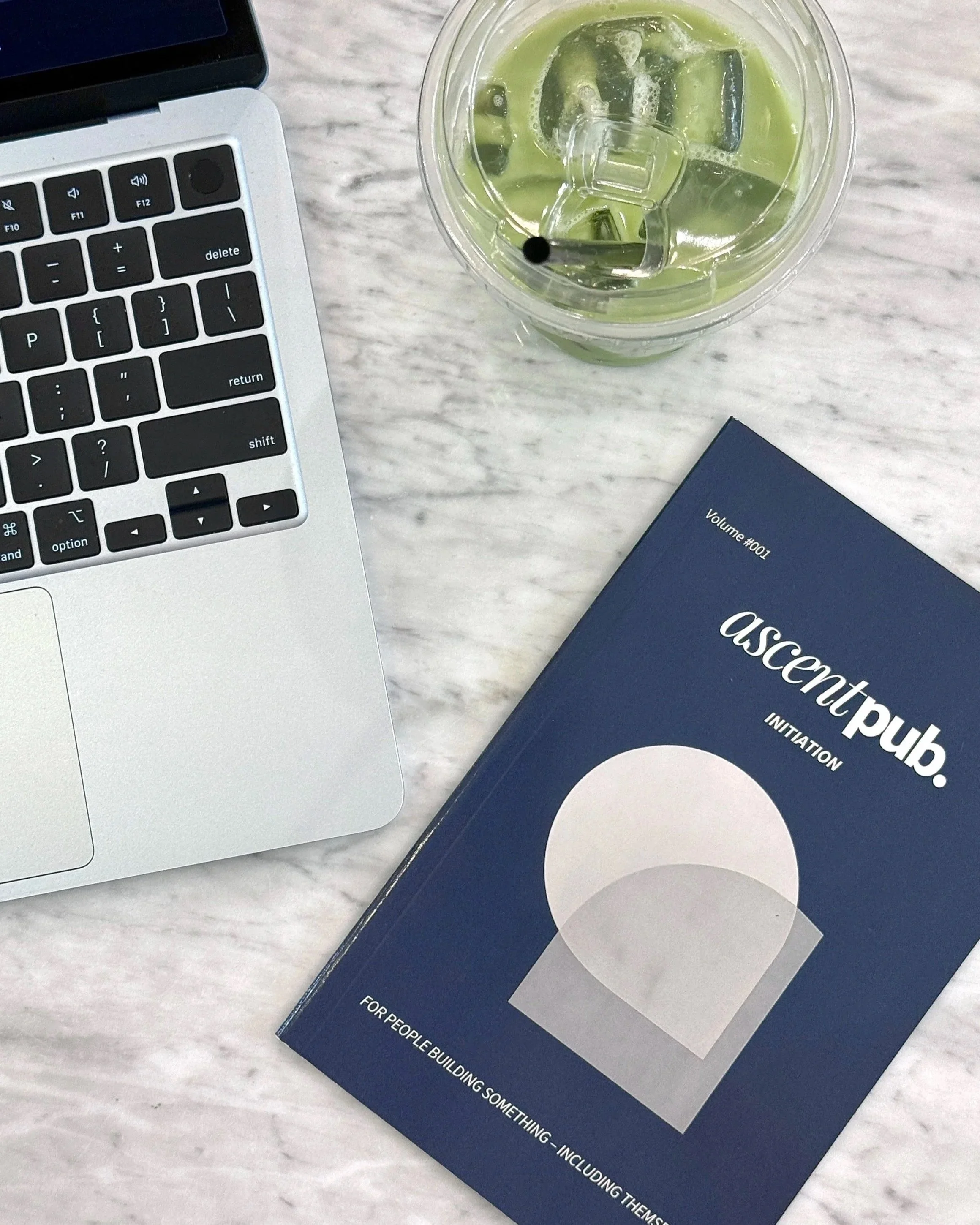 A laptop, a cup of green iced beverage with ice, and a navy blue book titled 'ascentpub' on a marble table.