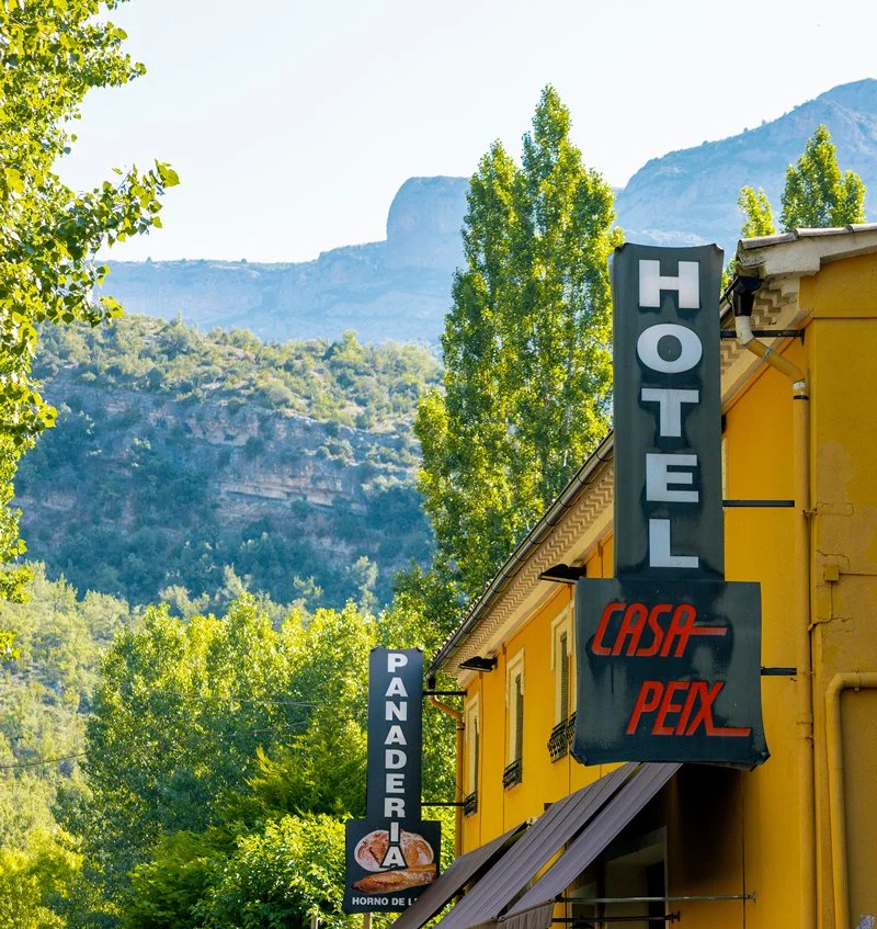 Letrero de un hotel y una panadería en un edificio amarillo, con montañas y árboles verdes en el fondo.