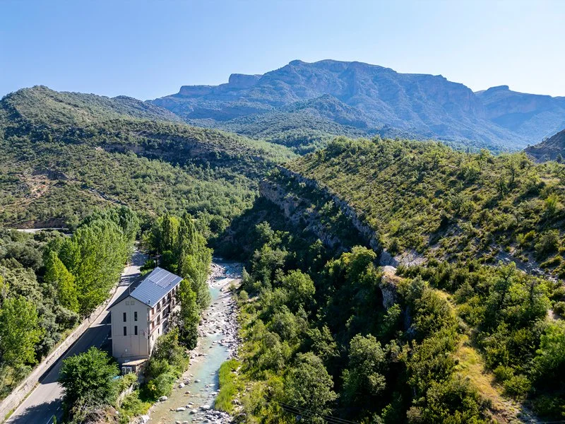 Paisaje de montañas verdes, río y edificio cerca de árboles en un día soleado.