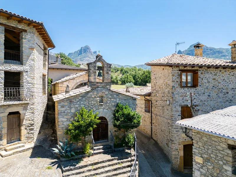 Imagen de un pueblo de montaña con casas de piedra, un pequeño templo con campanario y montañas al fondo bajo un cielo despejado.