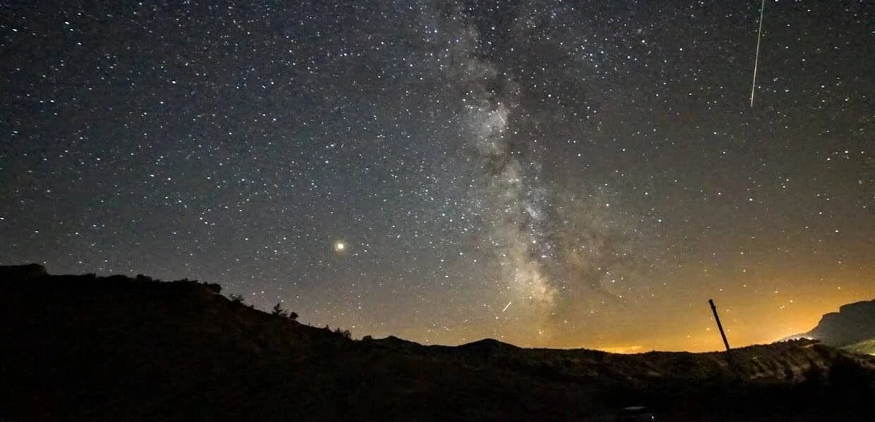 Cielo estrellado con la Vía Láctea visible en una noche despejada sobre un paisaje montañoso oscuro.