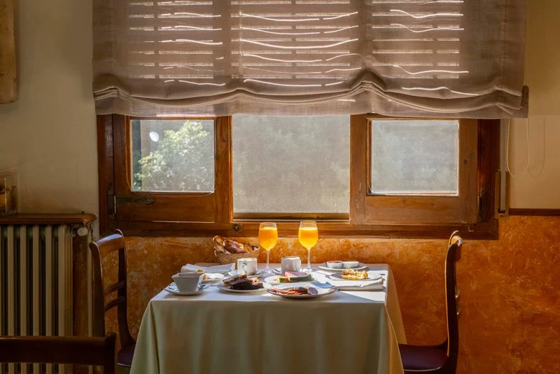 Mesa de desayuno con dos copas de jugo, platos con comida, tazas y utensilios, frente a una ventana con cortina