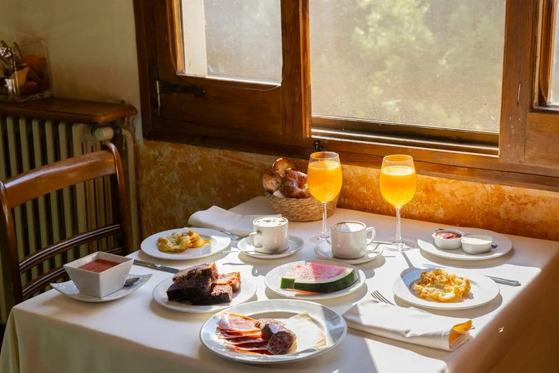 Mesa con desayuno que incluye jugo de naranja, café, pan, fruta, queso, y embutidos en una habitación con ventana
