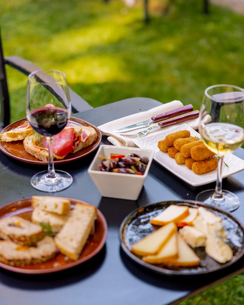 Mesa con comida, incluyendo emparedados, queso, aceitunas, croquetas, y dos copas de vino, en un ambiente al aire libre.
