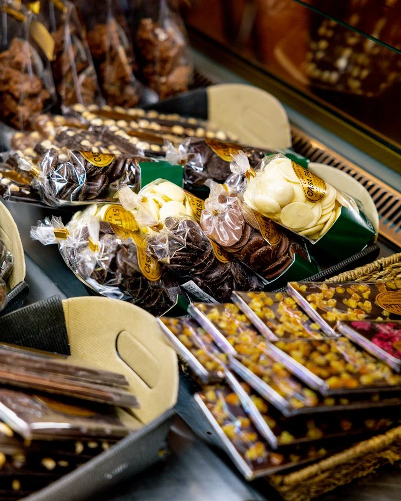 Variedad de chocolates y dulces en una tienda, empaquetados en bolsas transparentes y cajas, en estantes y bandejas.