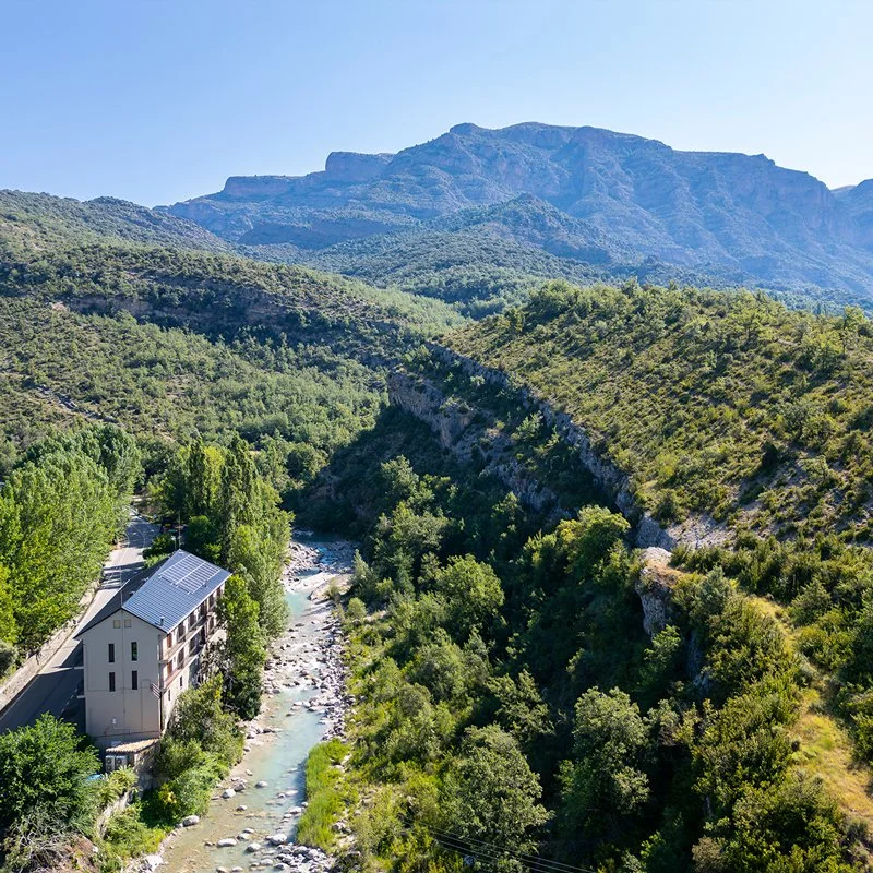 Paisaje montañoso con valle y río, casa junto al río, rodeado de árboles verdes bajo un cielo despejado.