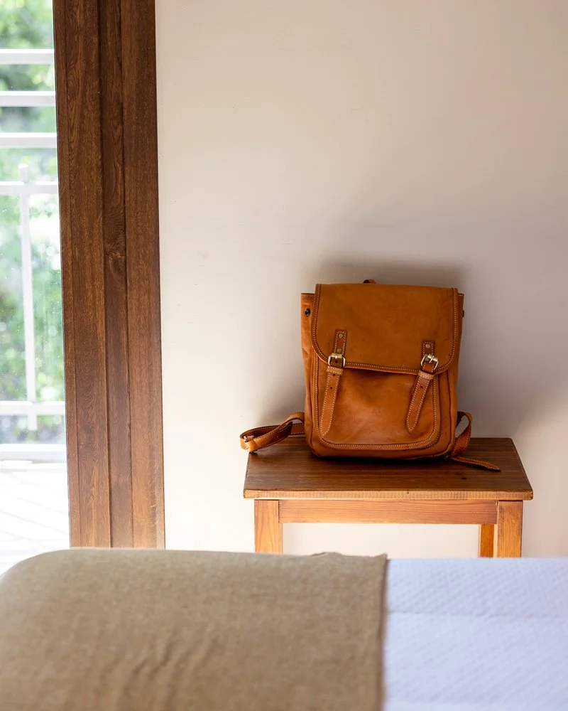 Una mochila de color marrón sobre una mesa de madera en una habitación con pared blanca y una ventana con marco de madera a la izquierda.