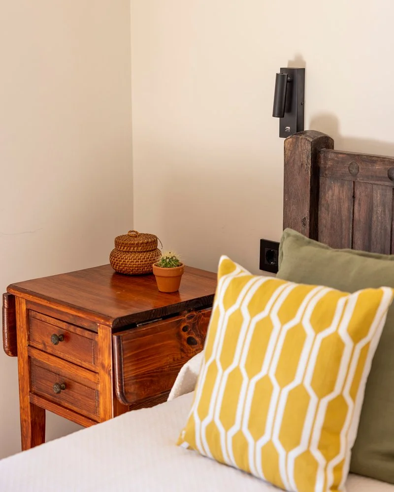 Detalle de una habitación con cabecera de madera, una cama con almohadas verdes y amarillas, y una mesa de noche de madera con una maceta y un cesto de fibras.