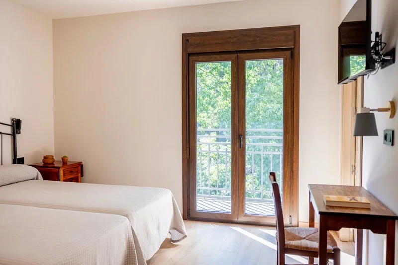 Habitación con cama doble, mesita de noche, balcón con vista a árboles, televisión y escritorio.