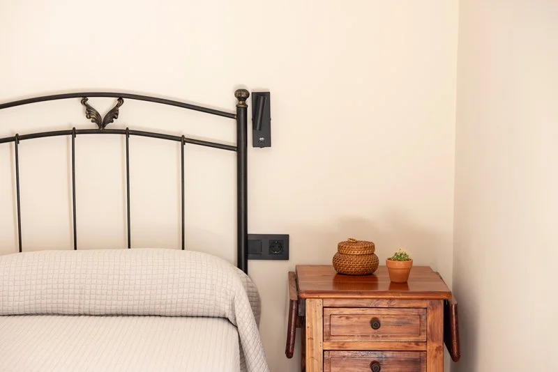 Parte de una cama con cabecera de hierro negro y una mesita de noche de madera con decoraciones de cestería y una planta en una maceta.