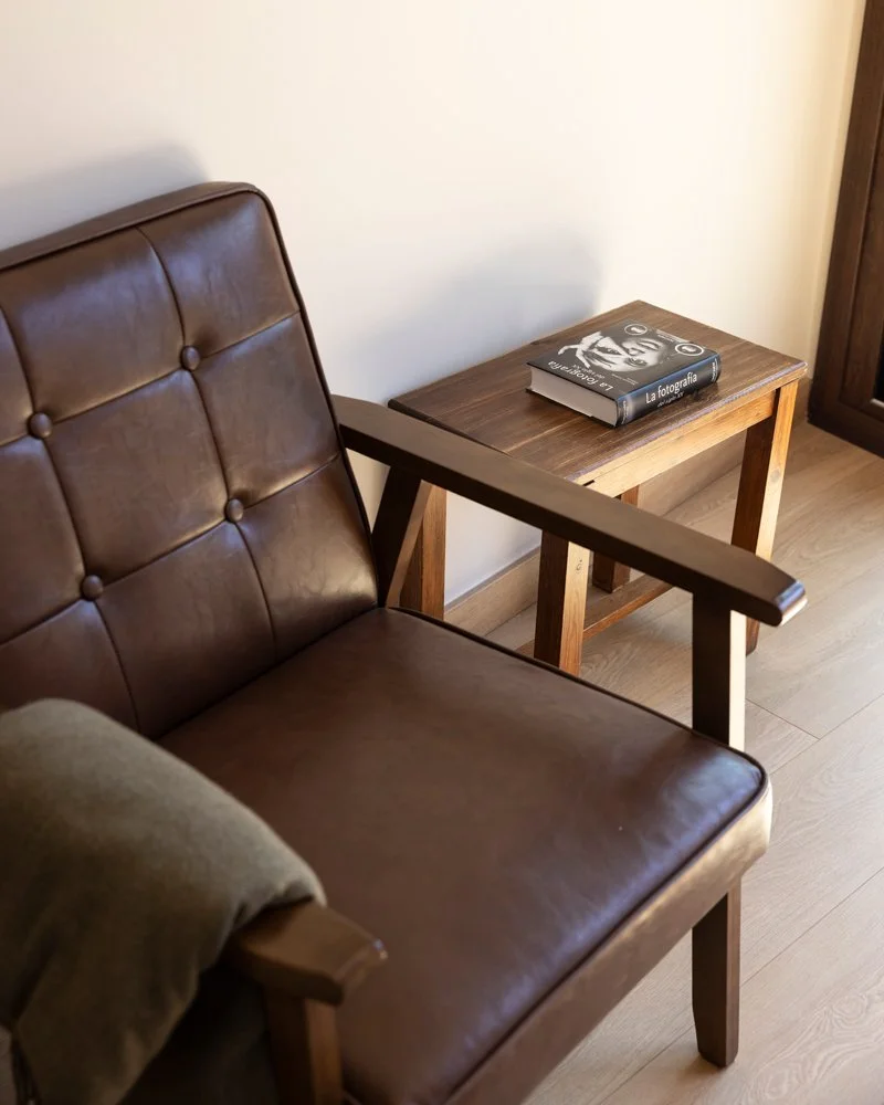 Sillón de color marrón con cojín gris y una mesa de madera con un libro titulado 'La fotografía' sobre ella en una habitación con suelo de madera.