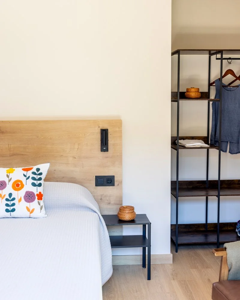 Habitación moderna con cama con colcha blanca, cojín con diseño floral, mesita de noche negra con cesta de mimbre, estantería de metal con ropa y accesorios, y suelo de madera clara.