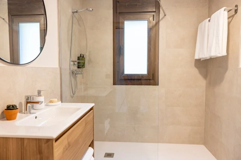  baño con paredes de baldosas beige, un espejo redondo, un lavabo de madera con artículos de tocador, una ventana de madera y un área de ducha con una barra de ducha y una estantería con productos, toallas blancas colgadas.