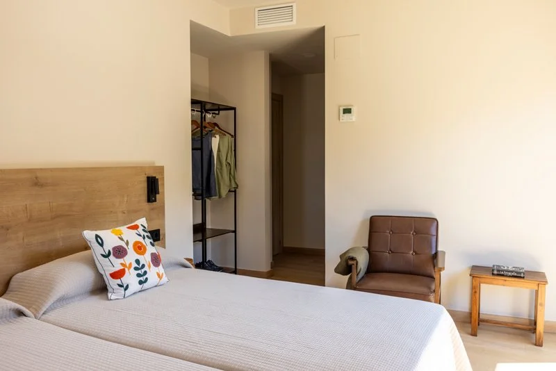 Habitación con cama, almohada decorativa, silla de cuero, mesa de madera y perchero abierto, paredes blancas, decoración sencilla.