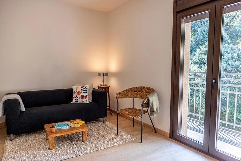 Habitación con cama, almohada decorativa, silla de cuero, mesa de madera y perchero abierto, paredes blancas, decoración sencilla.