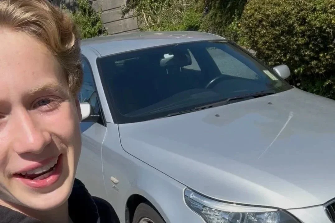 Close-up of a young man with light skin and blond hair smiling in front of a silver car, with trees and a wall in the background.