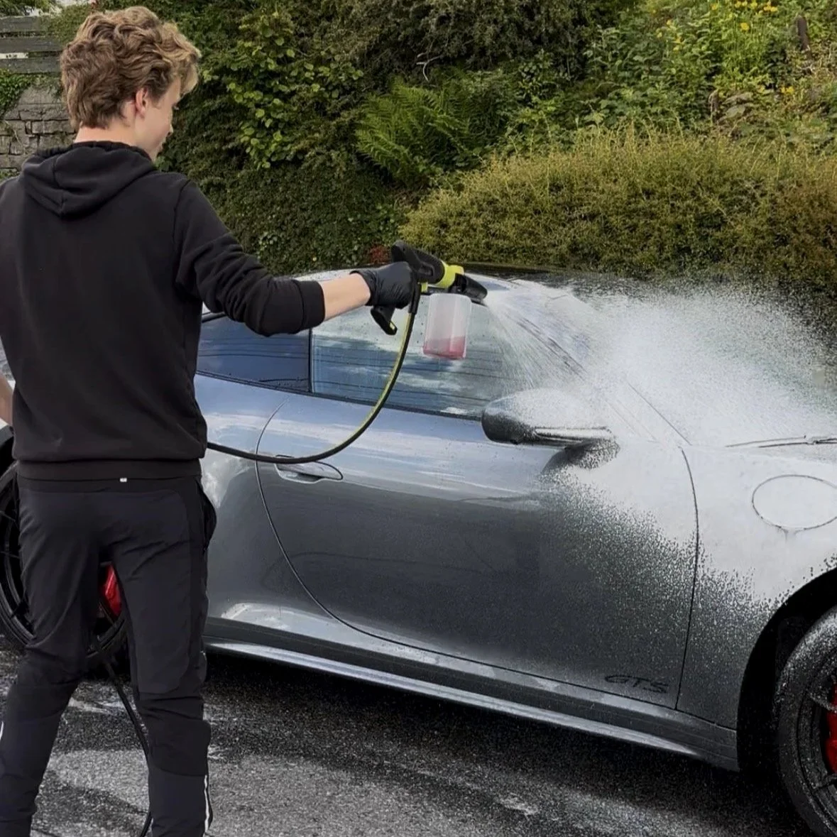A person in a black hoodie and black pants is pressure-washing a silver sports car outdoors, with a garden and greenery in the background.