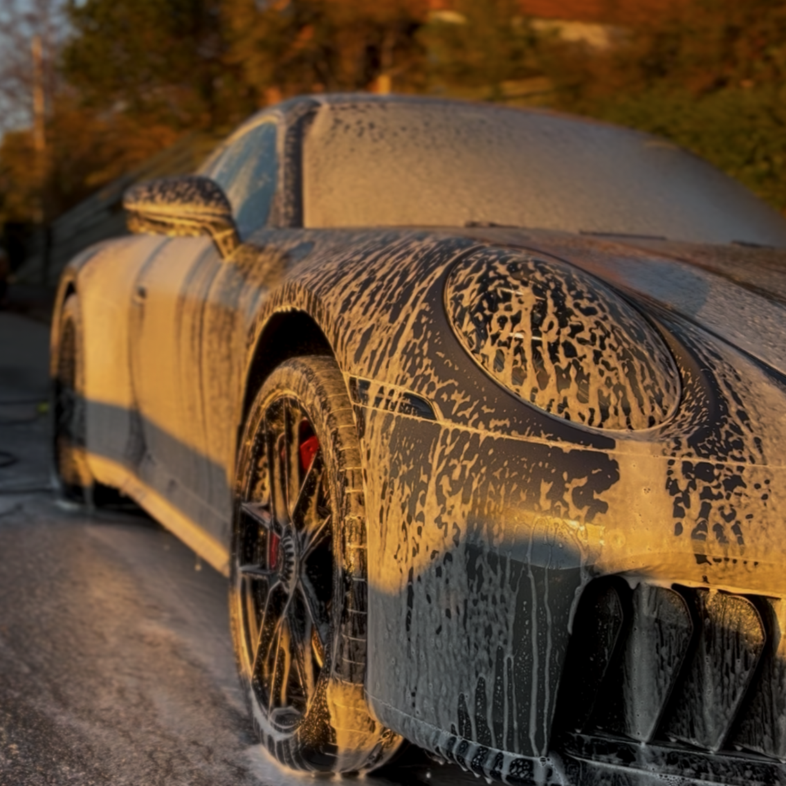 A luxury sports car covered in soap suds during a wash, with a foam pattern on the front rounded headlight and tire, at sunset.