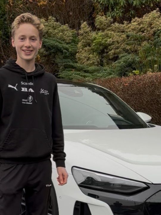 A young person standing next to a white car outdoors with lush green bushes in the background.
