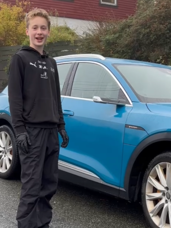 Young man standing next to a blue SUV on a driveway, smiling, with trees and a house in the background.