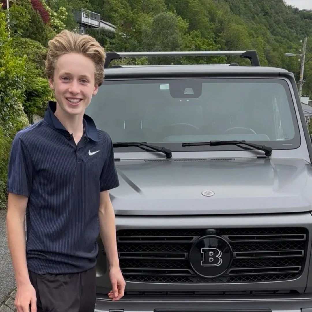 A young man with blond hair smiling and wearing a dark blue Nike polo shirt, standing next to a grey vehicle with a black grille and a large letter 'B' emblem on the front.