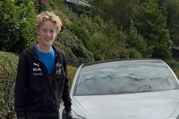 A young woman with curly blond hair, wearing a black jacket and blue shirt, standing next to a white car outdoors with trees and greenery in the background.
