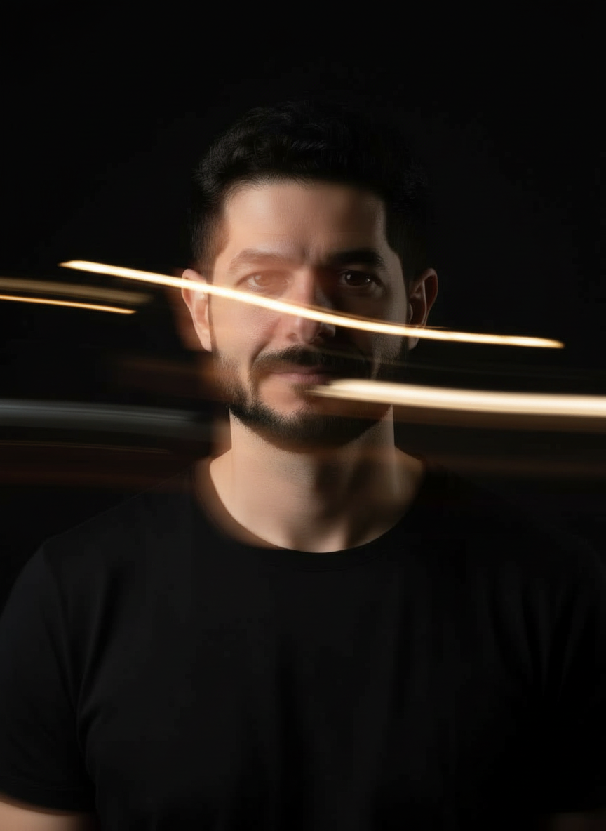 Portrait of AJ barber (Ahmad Jabateh) with dark hair and beard, wearing a black shirt, with light streaks across his face against a black background.